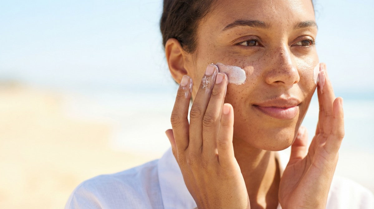 Close up happy young woman with natural freckles applying sunblock lotion to cheek outdoors during summer vacation under bright blue sky
